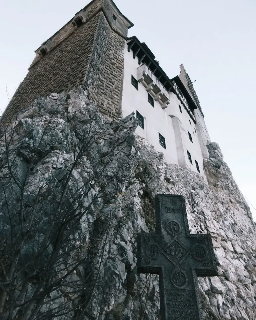 Bran Castle rising on its rock seen from below, with a stone cross in front