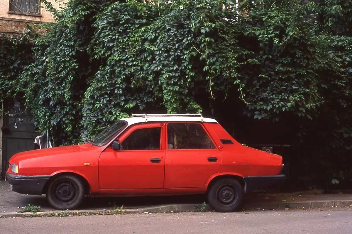Red vintage Dacia car parked on a street with ivy-covered walls