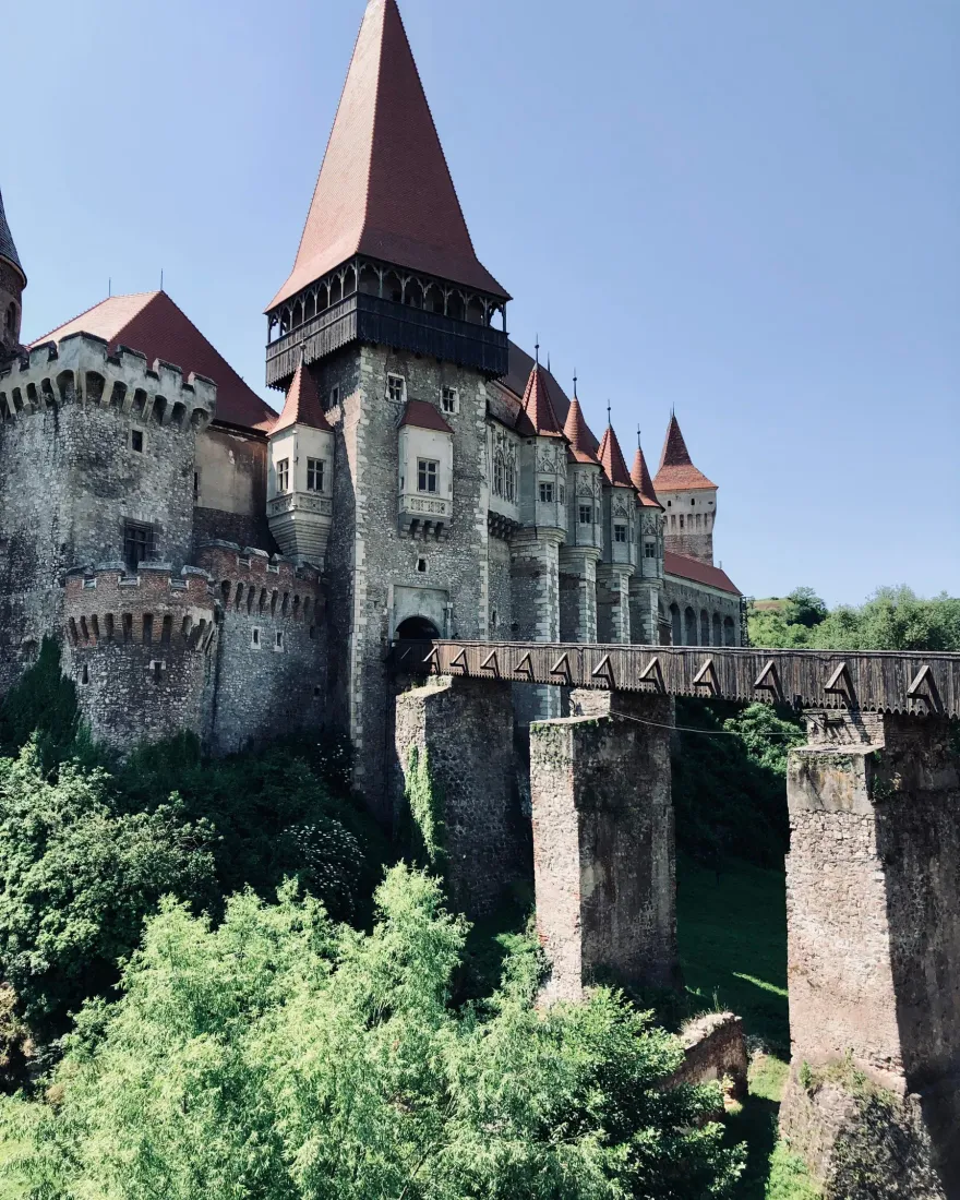 Side view of Corvin Castle above green trees, with its covered bridge and stone pillars