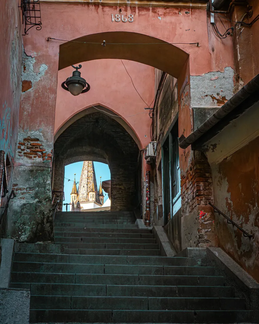 A vaulted passage and stairs in the old town of Sibiu with a church spire behind