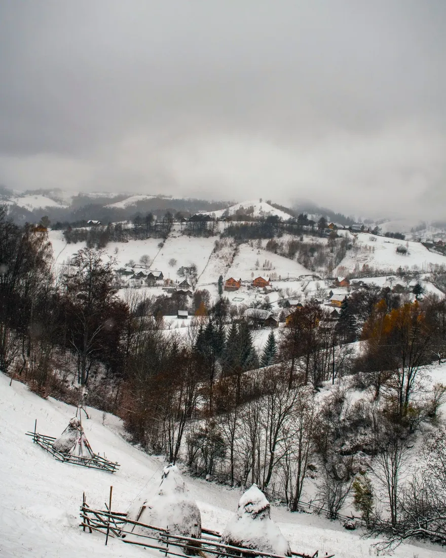 A snowy mountain village in Romania with haystacks and wooden fences under a grey winter sky